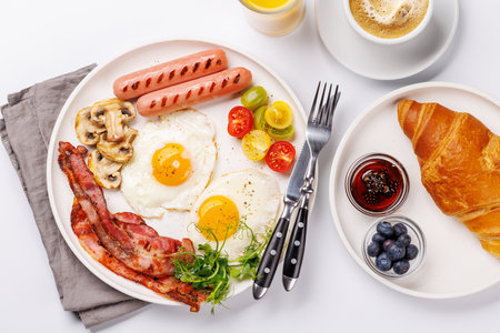 A classic breakfast plate featuring sunny-side-up eggs, crispy bacon, sausages, and fresh tomatoes, served with a hot cup of coffee for a hearty start to the dayの写真素材