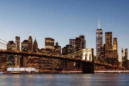 A panoramic view of the Brooklyn Bridge stretching towards Manhattan, with skyscrapers illuminated in warm hues against the fading skyの写真素材
