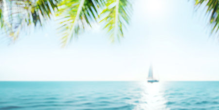 A serene seaside view with soft-focus palm leaves in the foreground and a sailboat gently gliding on the horizon under the warm sunlightの写真素材
