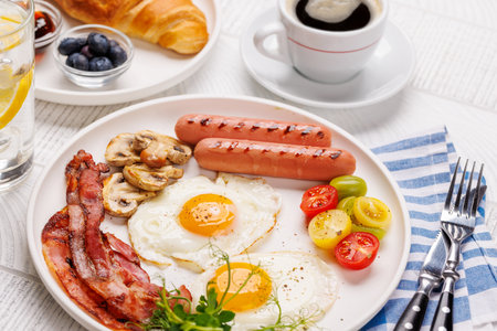 A classic breakfast plate featuring sunny-side-up eggs, crispy bacon, sausages, and fresh tomatoes, served with a hot cup of coffee for a hearty start to the dayの写真素材