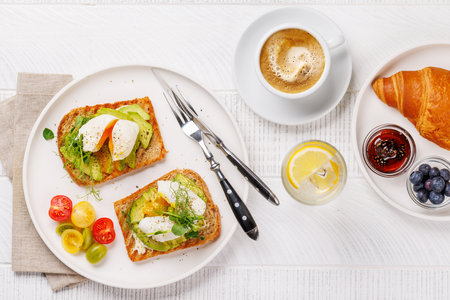 Breakfast with delicious avocado toasts topped with perfectly cooked poached eggs and crispy croissant with berries and jamの写真素材