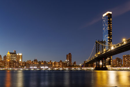 A panoramic sunset view of the Manhattan Bridge stretching towards Manhattan, with skyscrapers illuminated in warm hues against the fading skyの写真素材