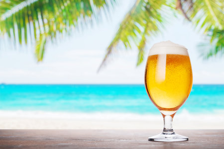 A glass of beer with a golden hue and frothy head, placed on a rustic wooden table with a beautiful summer sea landscape in the backgroundの写真素材
