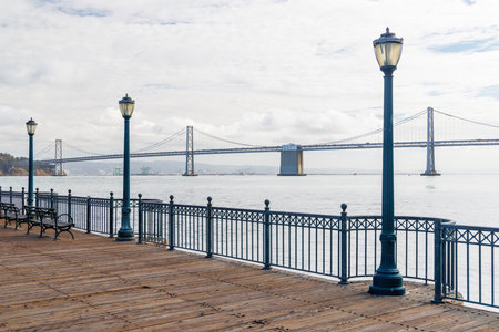 Scenic view of a classic wooden pier 7 stretching into the San Francisco Bay, framed by calm waters and Oakland Bay Bridge in the distance, under a bright skyの写真素材