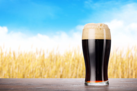 A glass of stout beer with a frothy head, placed on a rustic wooden table with a beautiful summer landscape in the backgroundの写真素材