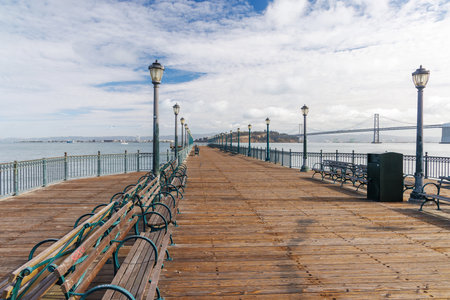 Scenic view of a classic wooden pier stretching into the San Francisco Bay, framed by calm waters and iconic city landmarks in the distance, under a bright skyの写真素材