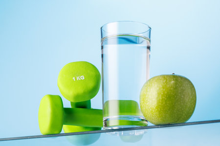 Glass of fresh water, green apple, and dumbbells on a glass table against a bright blue background, symbolizing fitness and healthの写真素材