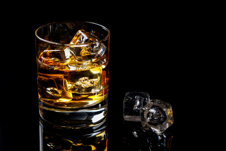 A refined glass of whiskey on the rocks, its amber hue glistening as the ice cubes slowly melt, set against a black background for a clean, elegant presentationの写真素材