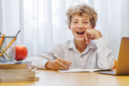 Schoolboy studying at home with laptop, notebook, and learning materials, smiling happily after finishing homeworkの写真素材