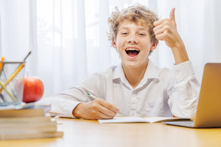 Schoolboy studying at home with laptop, notebook, and learning materials, smiling happily after finishing homeworkの写真素材