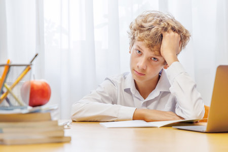 Schoolboy studying at home with laptop, notebook, and learning materials, looking thoughtful and...