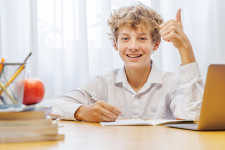 Schoolboy studying at home with laptop, notebook, and learning materials, smiling happily after finishing homeworkの写真素材