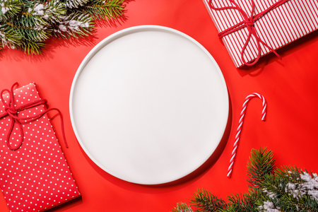 Empty plate with snowy fir branches and gift boxes on red background, Christmas food mockup. Flat layの写真素材