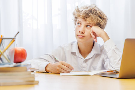 Schoolboy studying at home with laptop, notebook, and learning materials, looking thoughtful and...
