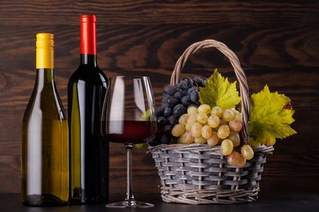 Red and white wine bottles with a glass and basket of fresh grapes on a table against a wooden wallの写真素材