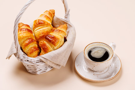 Aromatic croissants in wicker basket with cup of coffee on light background, cozy breakfast sceneの写真素材