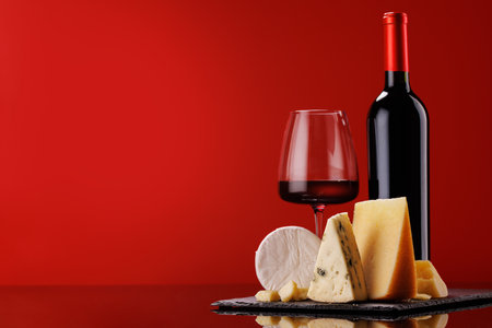 A refined selection of aged cheeses presented on a board, accompanied by a glass of wine, set against a red background. A perfect setting for a gourmet indulgenceの写真素材