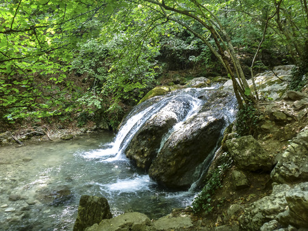 The small falls from transparent clear water, which flows down from big stones in the green deciduous forestの写真素材