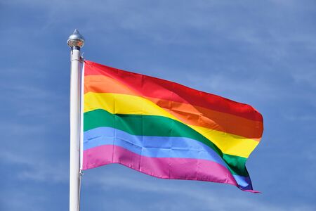 The pride flag on a white flagpole against a blue sky.