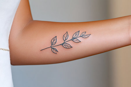 Minimalist tattoo of a delicate branch with leaves is displayed on a woman's forearmの素材