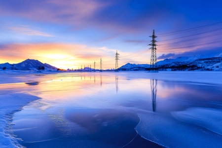 Stunning sunset over a frozen lake with power lines reflecting in the icy water and snow-capped mountains in the backgroundの素材