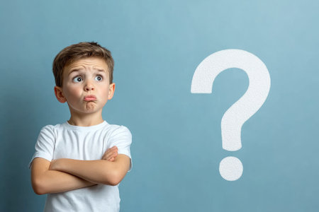 Pensive schoolboy thinking about a solution, looking up at a question mark on a blue background, arms crossedの素材