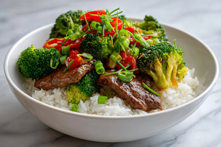 Beef and broccoli stir fry with red bell peppers and green onions is served over white rice in a white bowlの素材