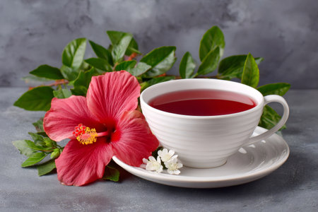 Hibiscus tea infusing in a white cup with a fresh flower and green foliageの素材