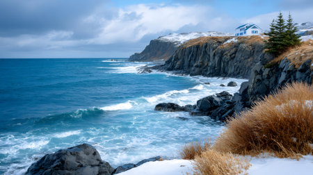Ocean waves crashing against rocky cliffs with a small white house on the snowy clifftopの素材