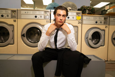 Young handsome man in shirt and tie sitting on washing machine in laundromatの写真素材
