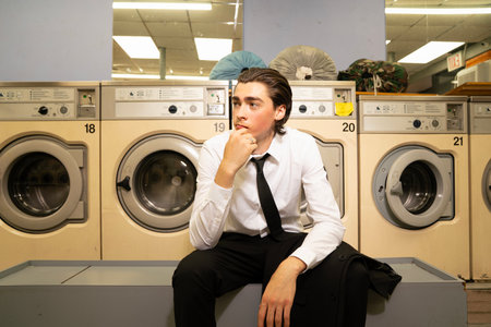 Young man in shirt and tie sitting in front of washing machine in laundromatの写真素材