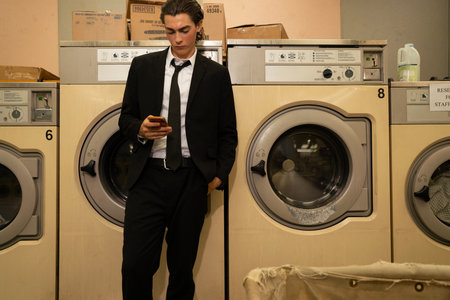 A young man in a black suit using a mobile phone in a laundromatの写真素材