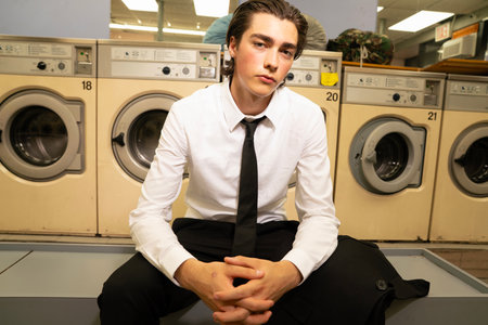 Young man in shirt and tie sitting in front of washing machine.の写真素材