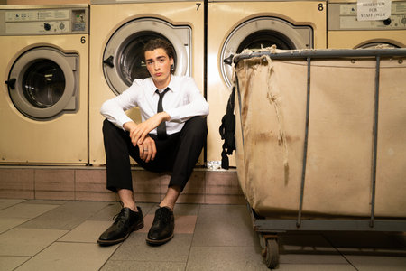 Young man in shirt and tie sitting in front of washing machine.の写真素材