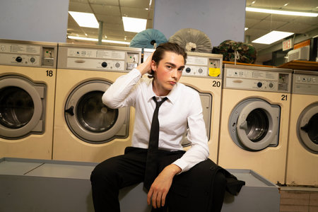 A young man in a shirt and tie sitting in front of washing machinesの写真素材