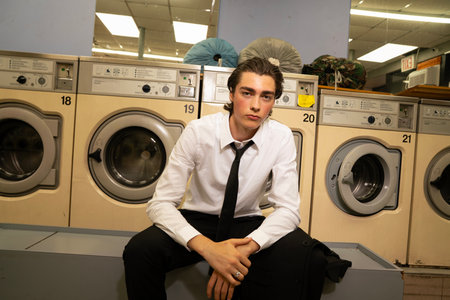 Young man in shirt and tie sitting in front of the washing machineの写真素材