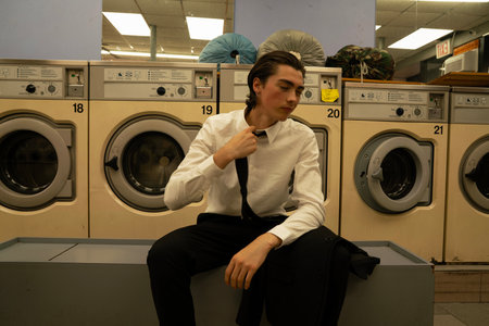 young man in shirt and tie sitting in front of washing machine in laundromatの写真素材