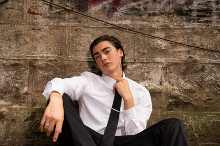 Young man in white shirt and black pants sitting on a stone wallの写真素材