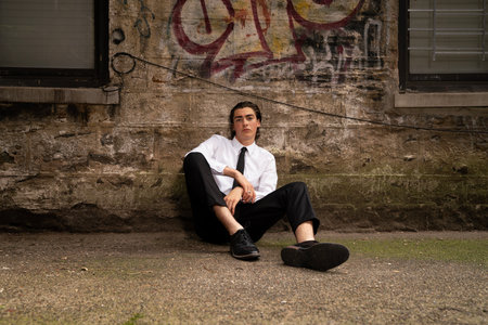 A young man in a white shirt and black trousers is sitting on the street near the old wall.の写真素材