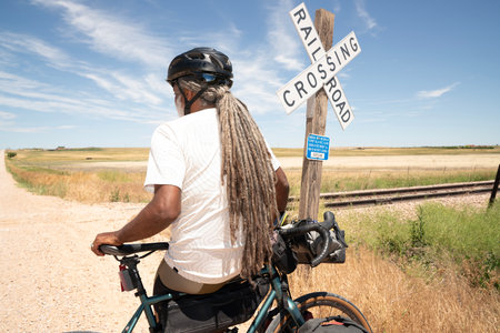 Rear view of a man with dreadlocks riding a bicycle along a railroad crossing.の写真素材