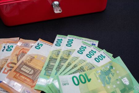 a lot of euro banknotes lie spread out in front of a red cash boxの写真素材