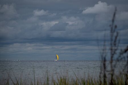 far on the sea floats a sailboat with a bright yellow sail and the sky is full of cloudsの写真素材