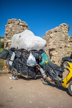 a scooter with a trailer is standing on a sandy path, full to bursting with garbage bagsの写真素材