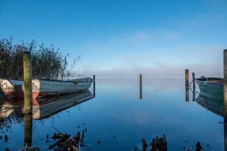 on a lake some rowboats are moored and on the lake there is fog and the lake is quite calmの写真素材
