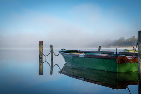 on a lake some rowboats are moored and on the lake there is fog and the lake is quite calmの写真素材
