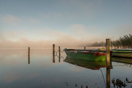on a lake some rowboats are moored and on the lake there is fog and the lake is quite calmの写真素材