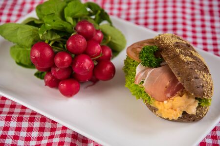 on a white plate lies a sliced bread roll filled with sausage, cheese and egg and as decoration a bunch of radishesの写真素材