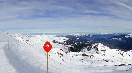 a red sign with the number 5 stands next to a snow covered ski route and the sky is blueの写真素材