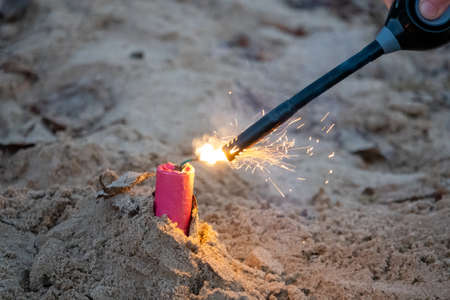 A red firework stuck in the sand is lit with a black stick lighterの写真素材