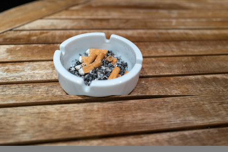 Cigarette butts lie crushed in a white ashtray standing on a wooden tableの写真素材